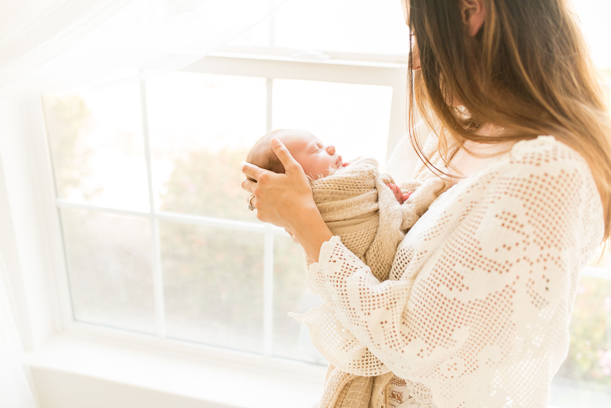 Mother gently strokes baby's head standing by the window captured by Sweet Beginnings Photography by Stephanie