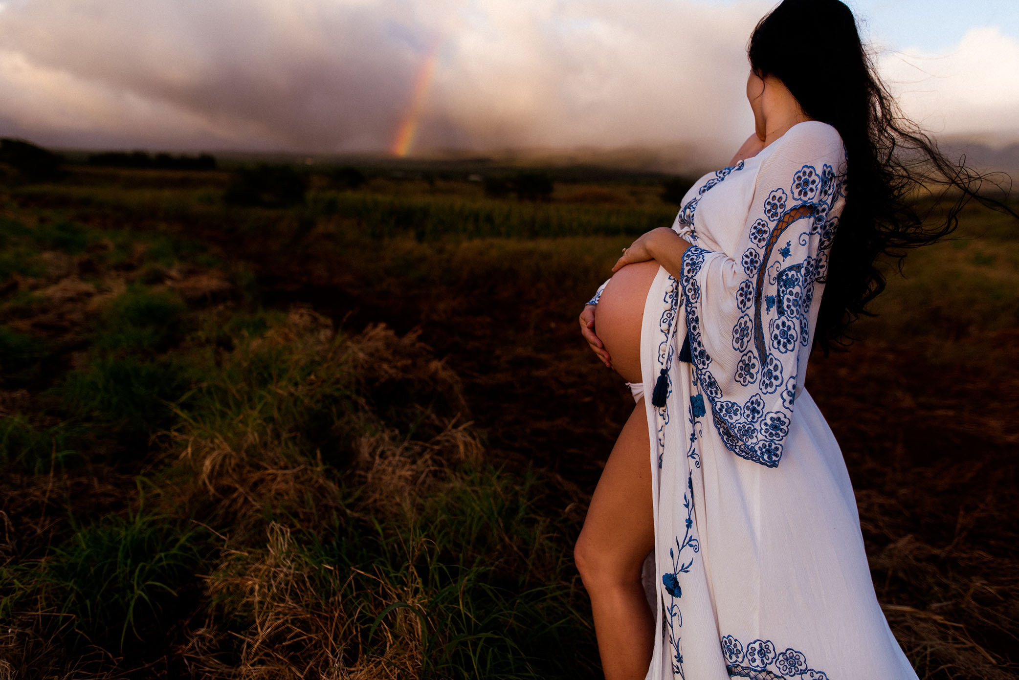 Pregnant mom holds her belly in Fillyboo dress looking at Maui rainbow captured by Sweet Beginnings Photography by Stephanie