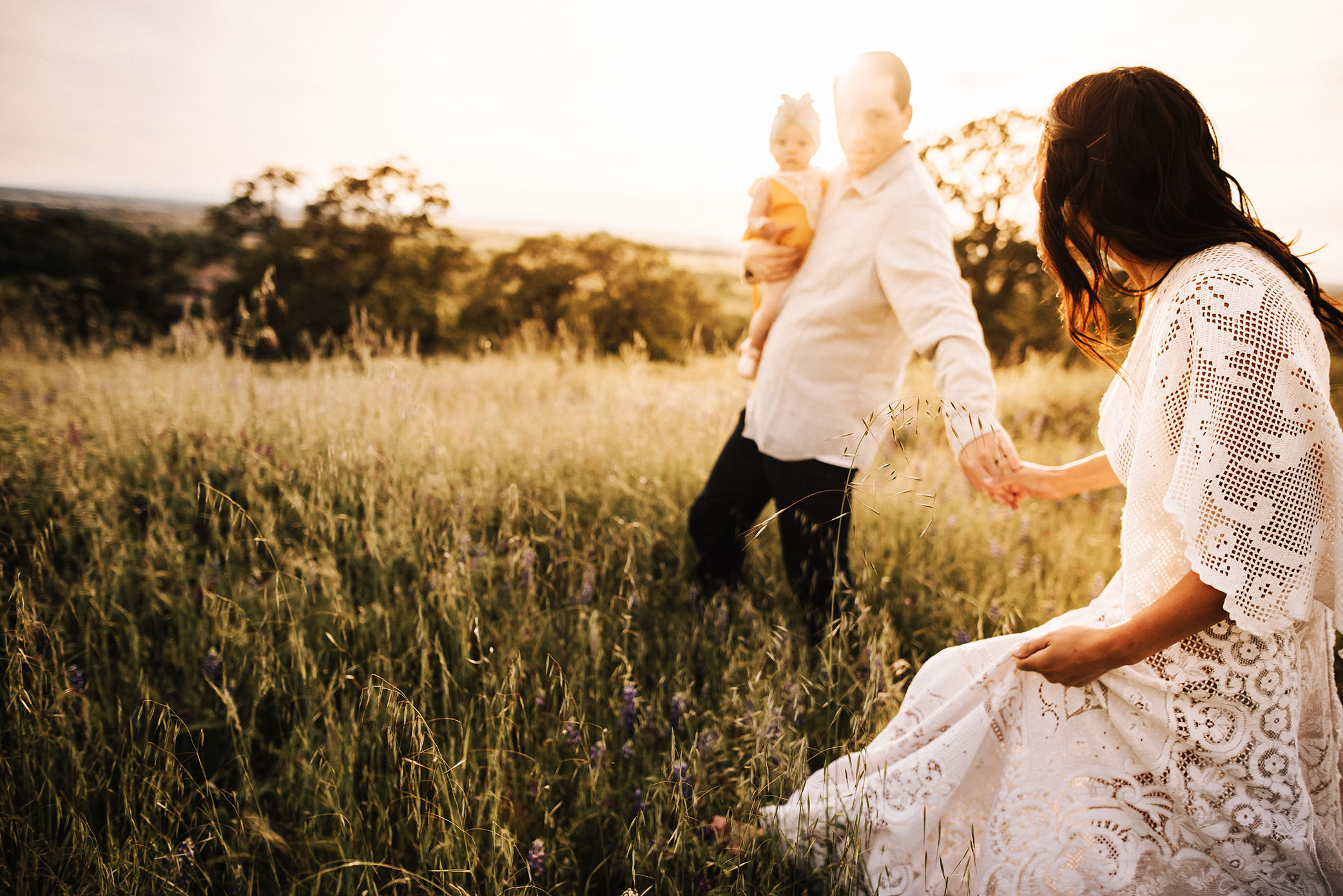 Mom and dad walk through a grassy hill with sun beaming in captured by Sweet Beginnings Photography by Stephanie 