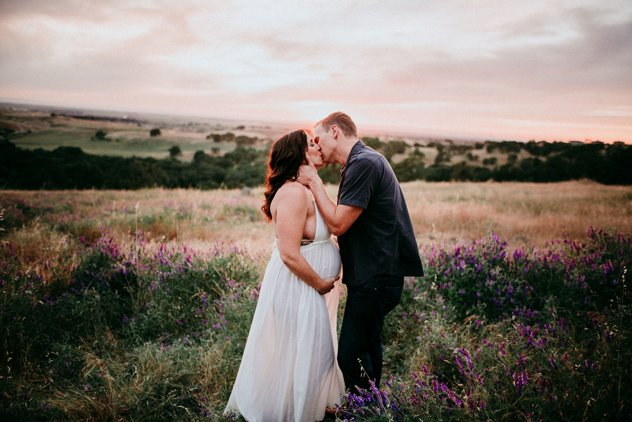 Pregnant woman kissing husband at sunset wearing white dress captured by Sweet Beginnings Photography by Stephanie