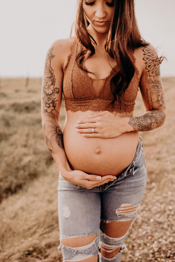 Mother expecting baby looks down and holds her belly wearing jeans and bralette