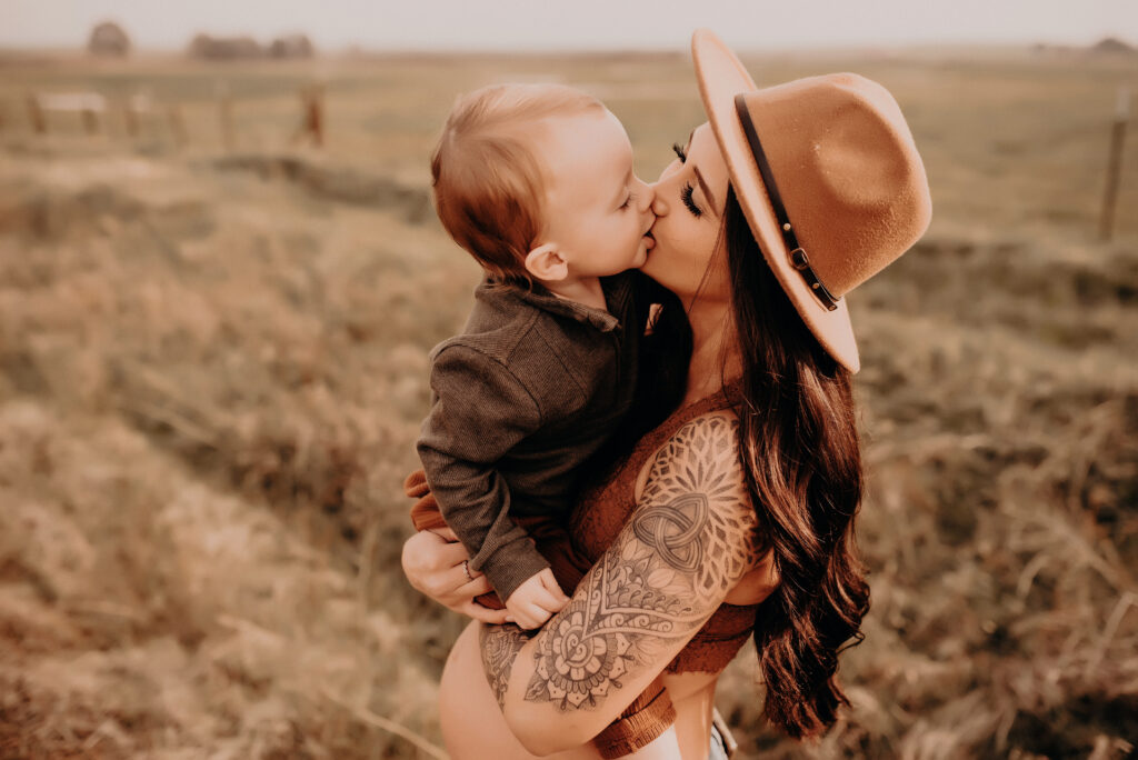Pregnant mama kisses her toddler while holding him above belly wearing ha, jeans and bralette
