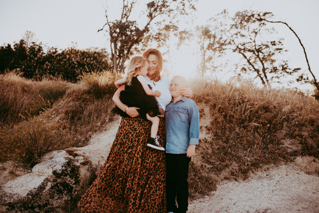 Mom wearing cheetah print maxi skirt holds her toddler wearing black tulle dress and arm around son wearing chambray button up